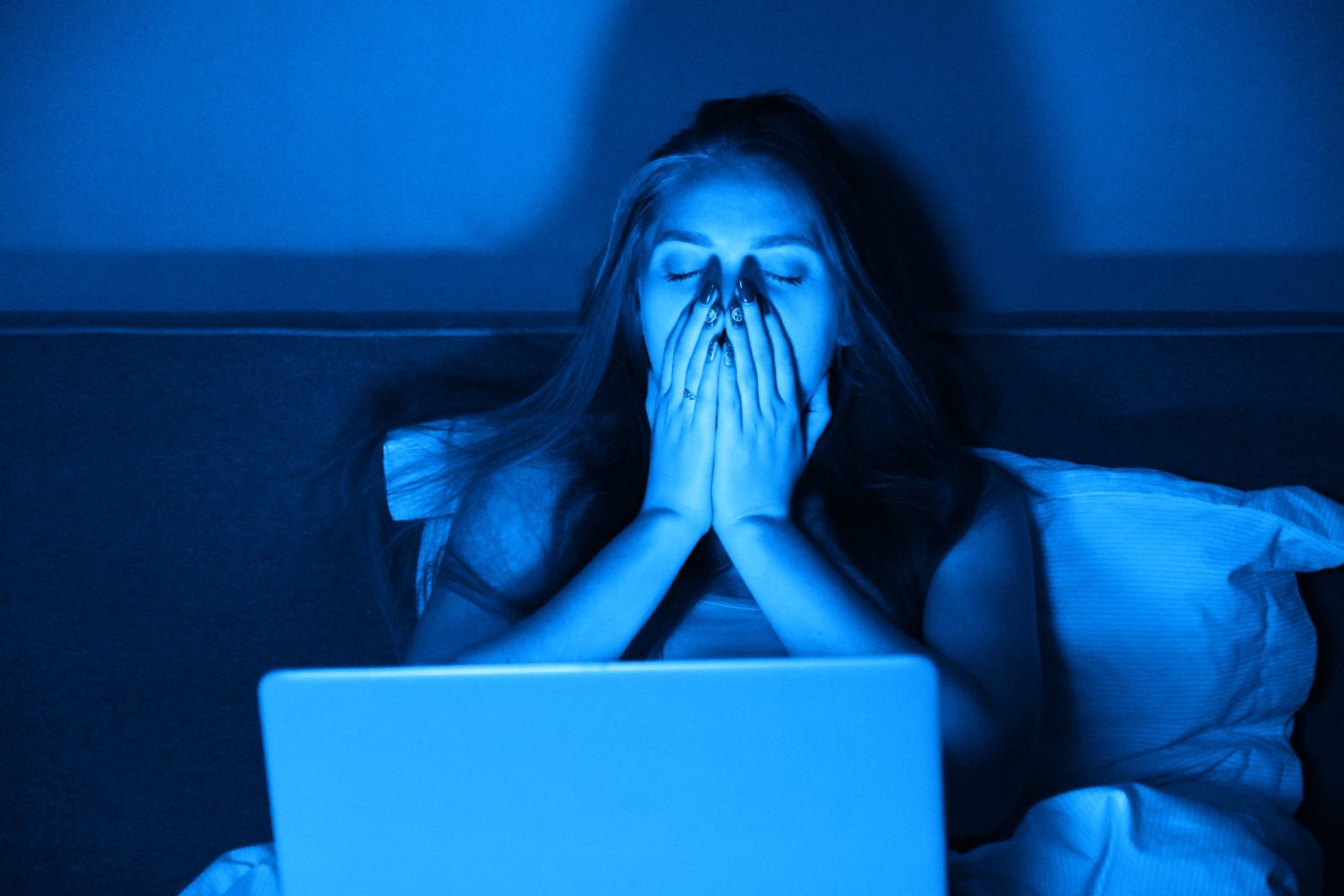 Femme sous le choc, les mains sur le visage, éclairée par son ordinateur la nuit